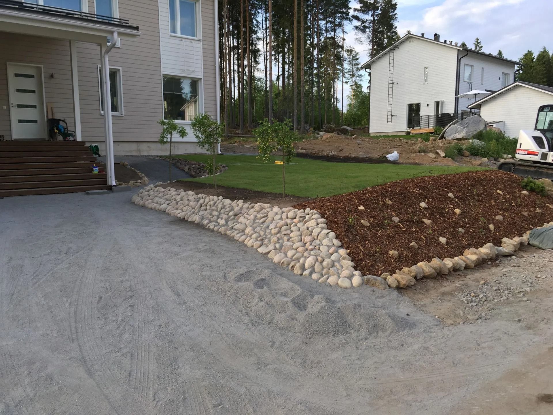 Maisemoitu etupiha, jossa kivinen tukimuuri, sorapäällysteinen ajotie ja taloja asuinalueella
