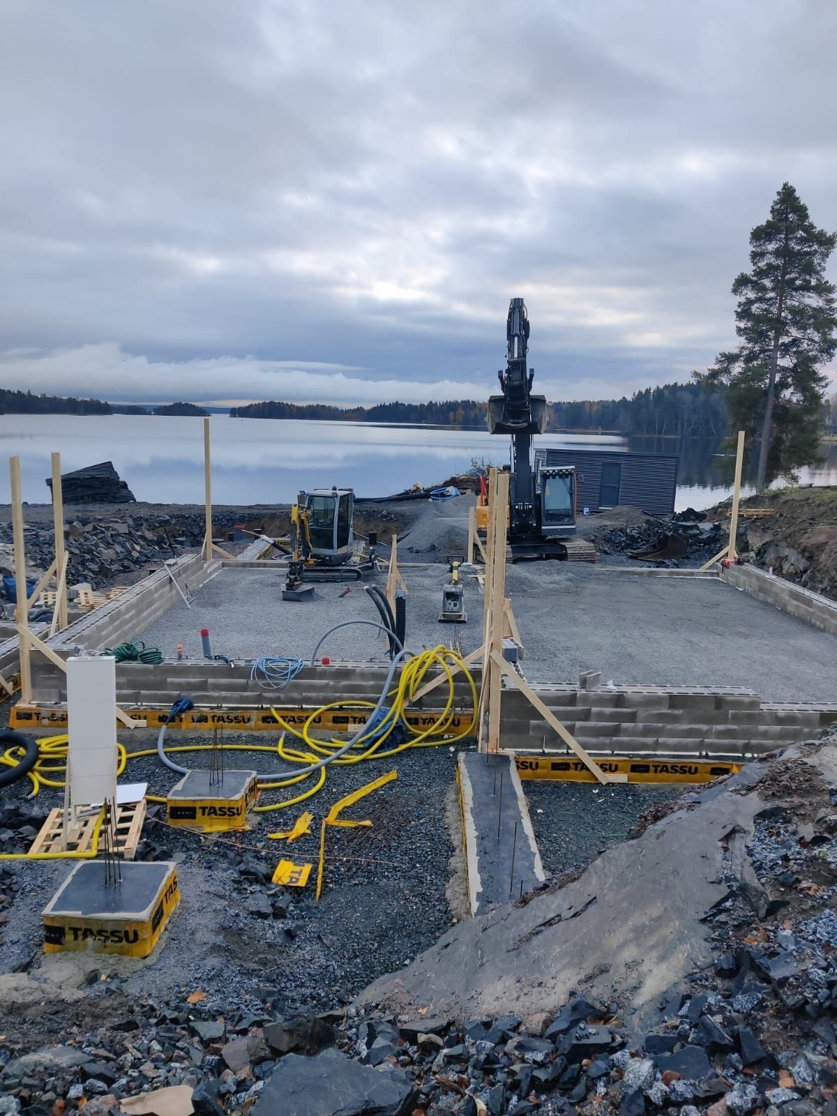 Rakennustyömaa mutaisella maalla, keltaisilla letkuilla ja järvenrannalla pilvisen taivaan alla