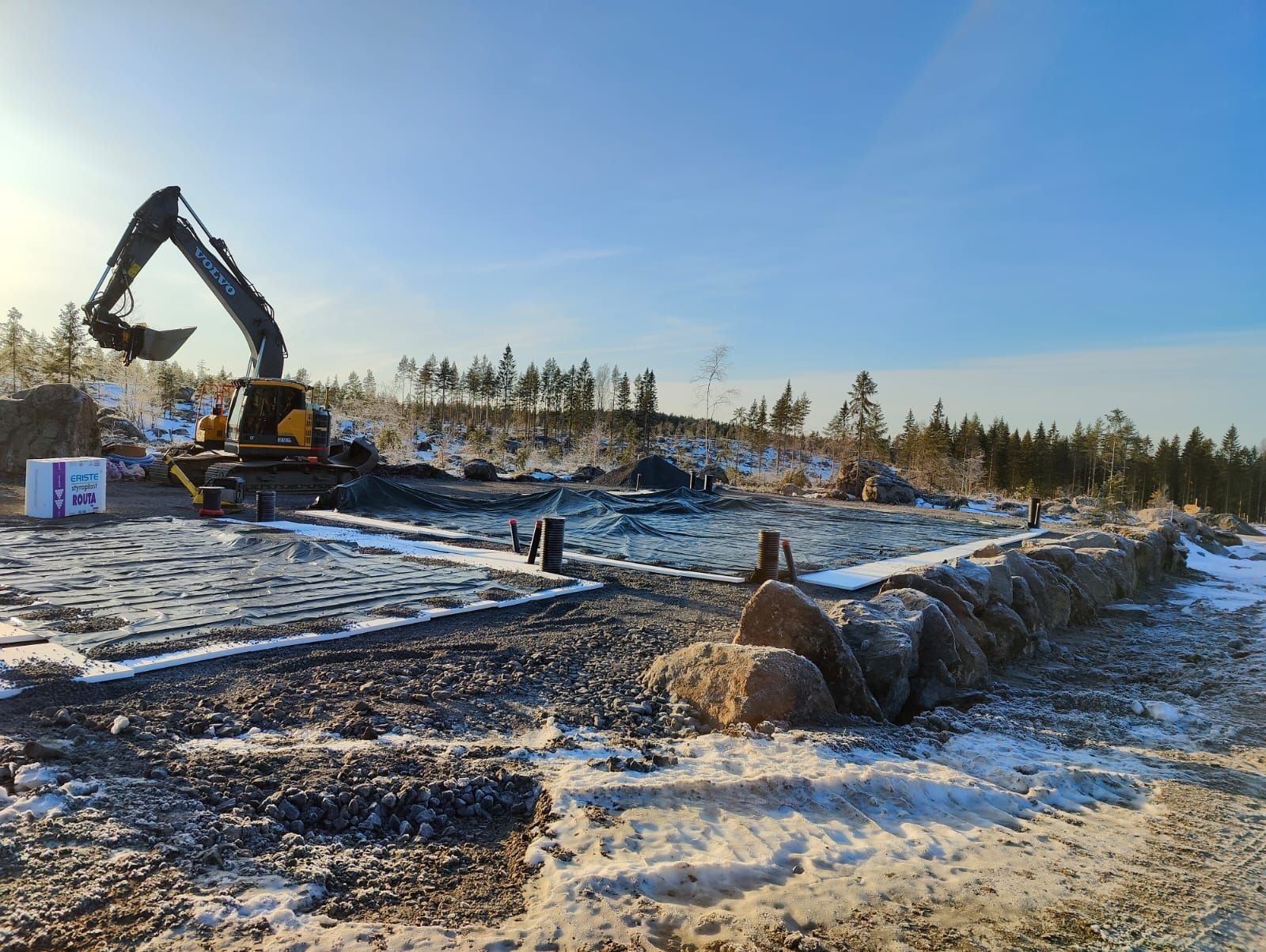 Kaivinkone työskentelee lumisella rakennustyömaalla sinisen taivaan alla, taustalla puita
