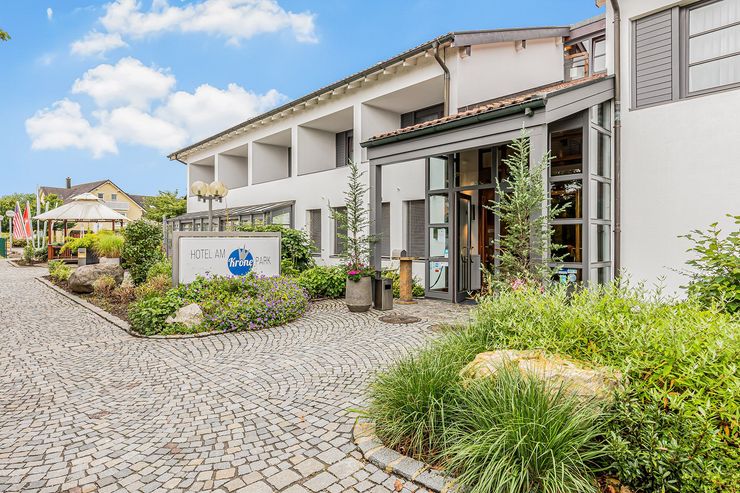 Hoteleingang des Hotel am Krone Park mit Kopfsteinpflasterweg, Schild, Glastüren und grüner Bepflanzung