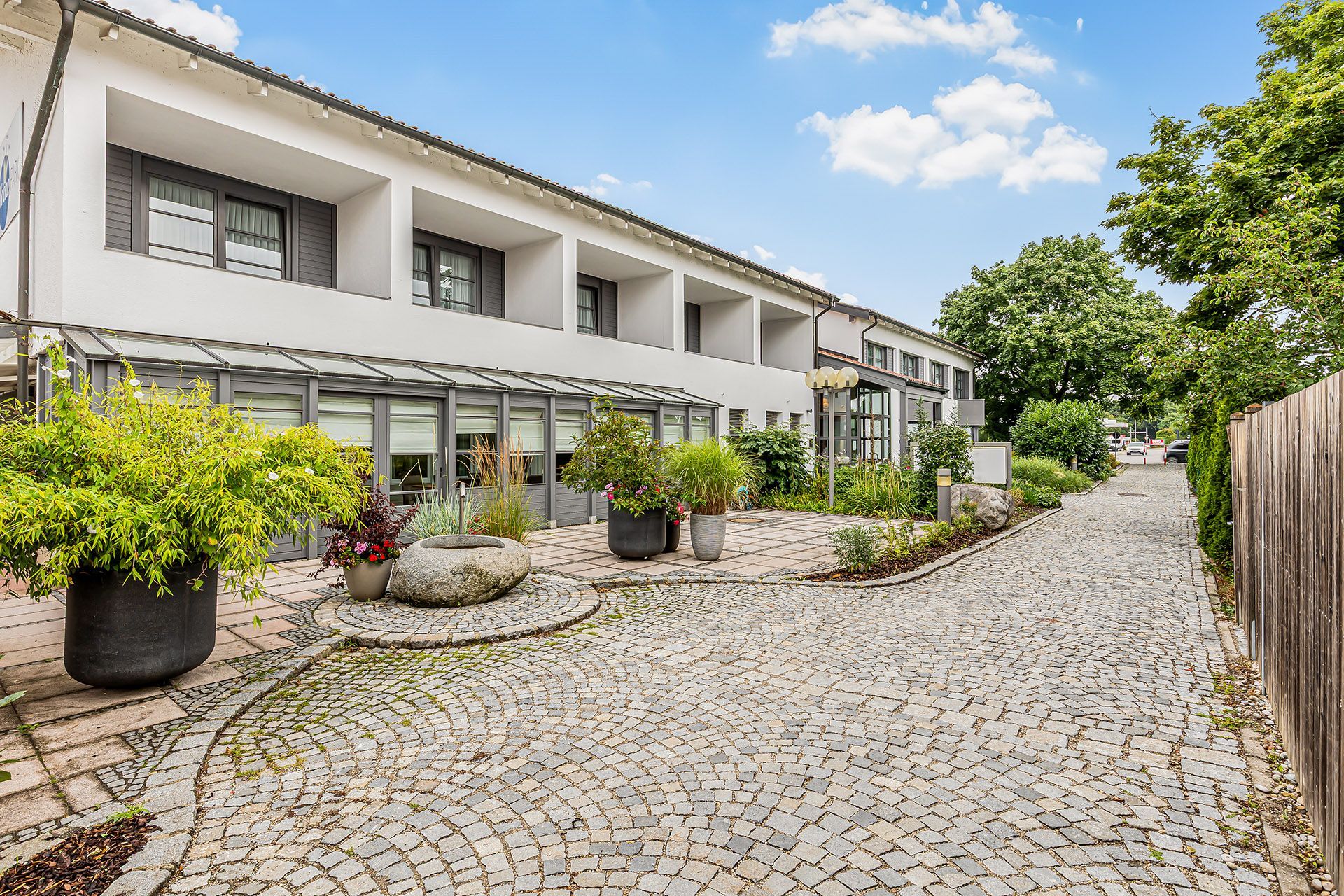 Außenansicht des Hotel am Krone Park mit Balkonen, üppigem Grün und einem Kopfsteinpflasterweg an einem sonnigen Tag.