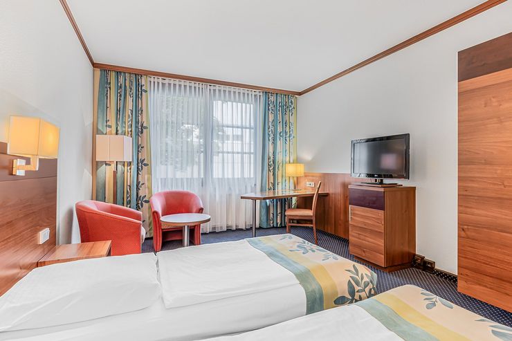 Doppelzimmer mit zwei Einzelbetten, roten Sesseln, Schreibtisch, Fernseher.