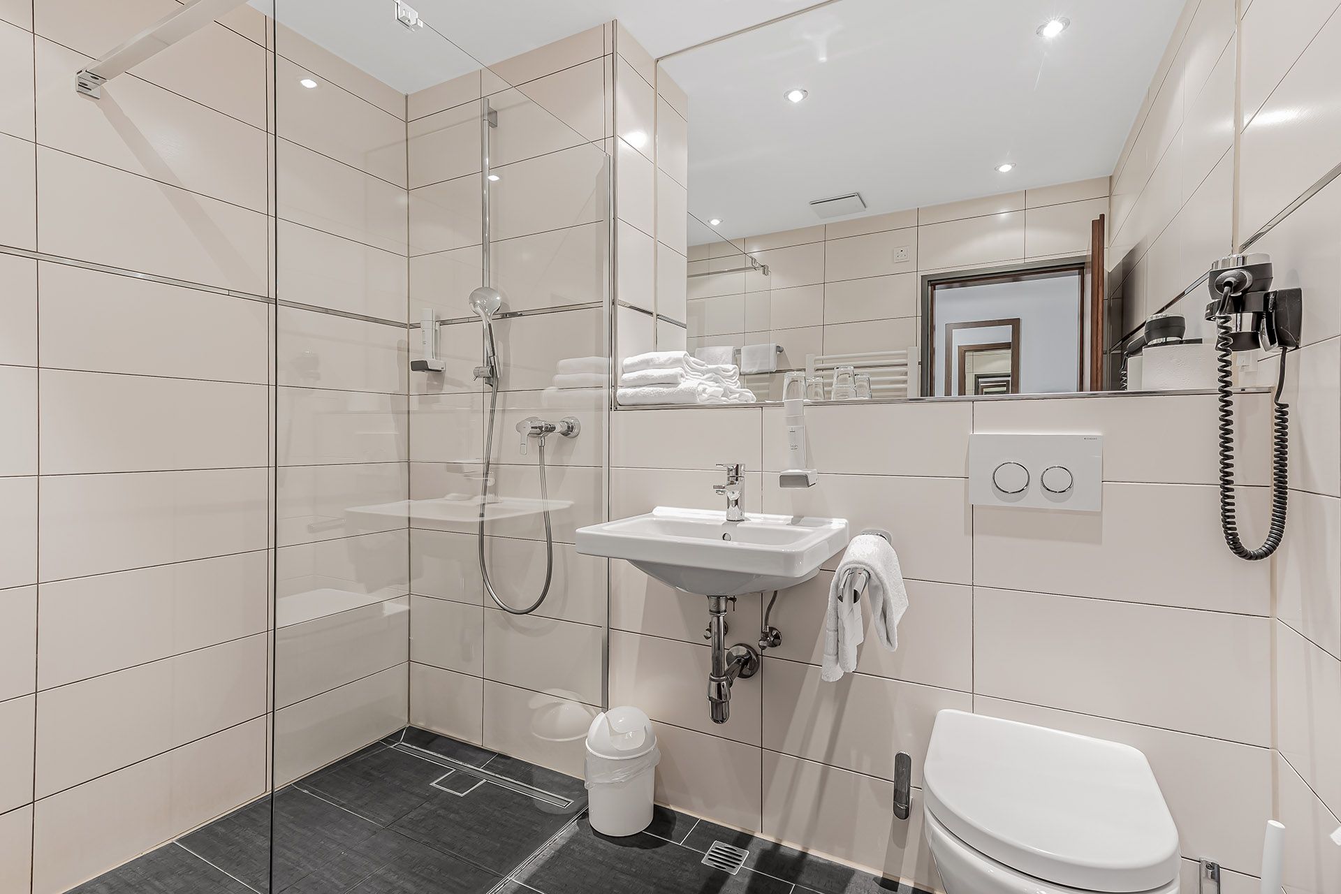 Modernes Hotelbadezimmer mit weiß gefliesten Wänden, Dusche, Waschbecken, Toilette und Spiegel.