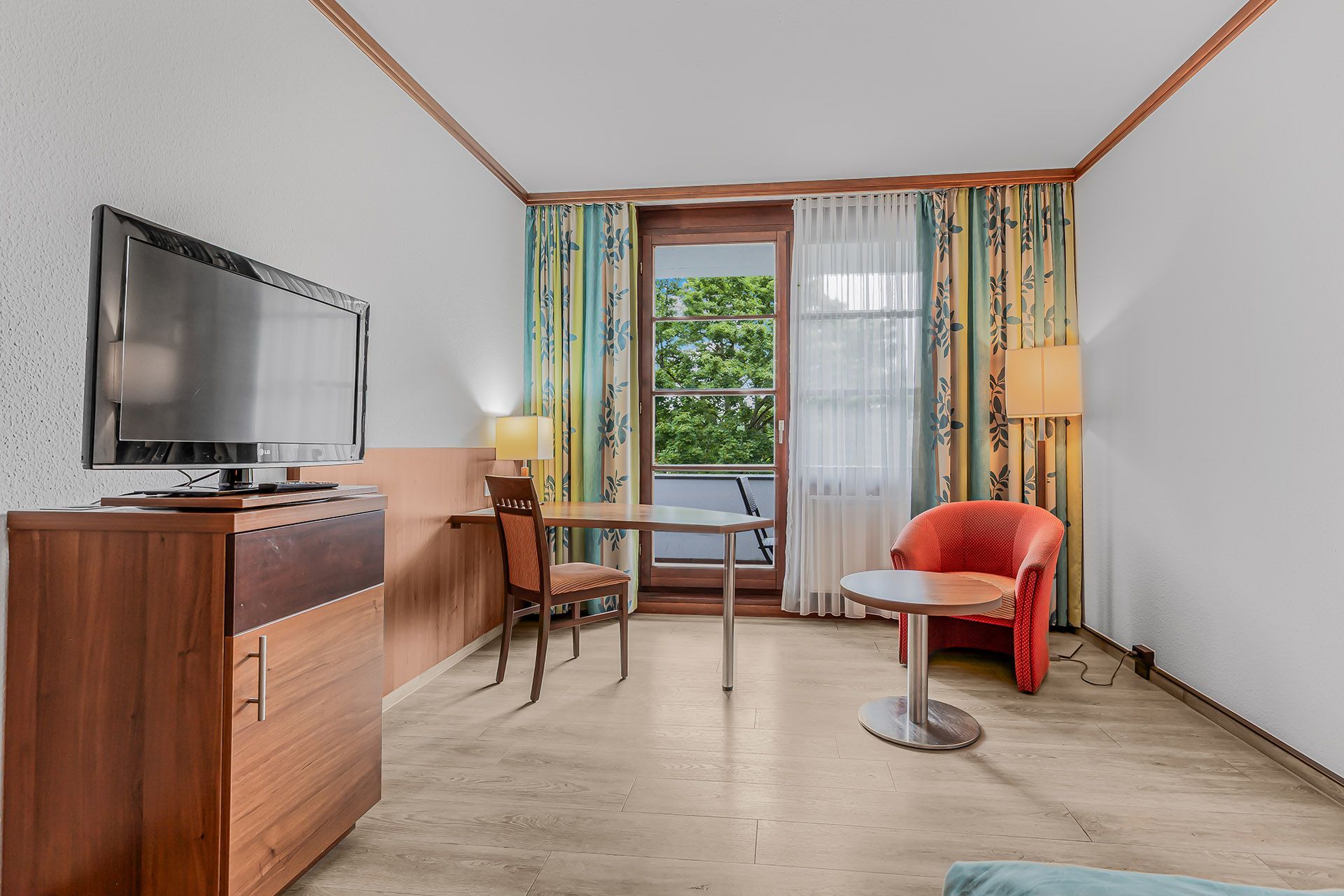 Hotelzimmer mit Fernseher, Schreibtisch, Stuhl, rotem Sessel und Balkontüren.