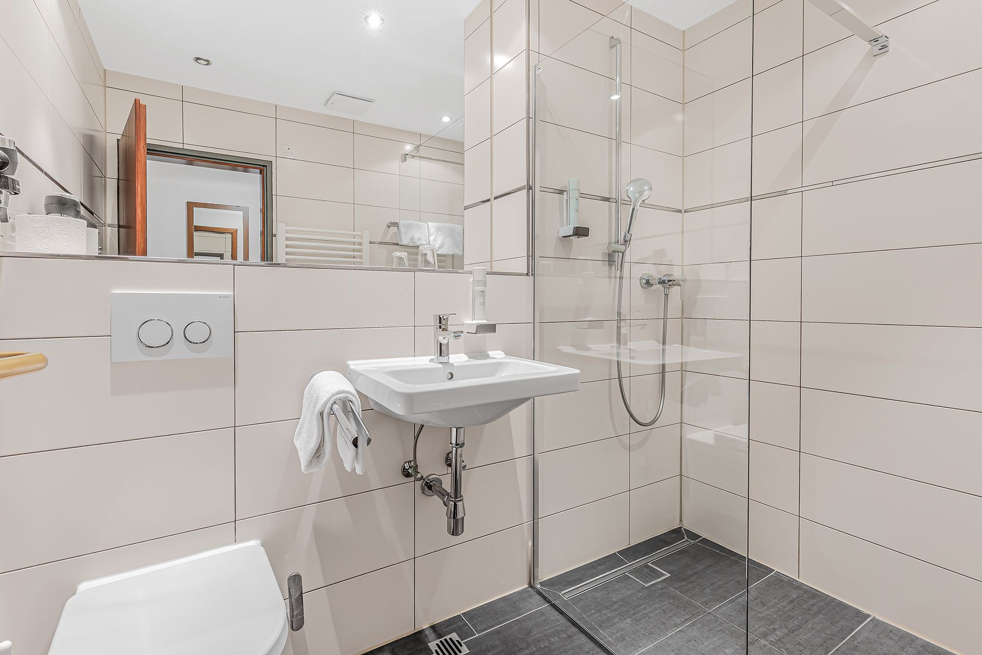 Modernes Badezimmer mit Glasdusche, Waschbecken, Toilette und hellbeige gefliesten Wänden.