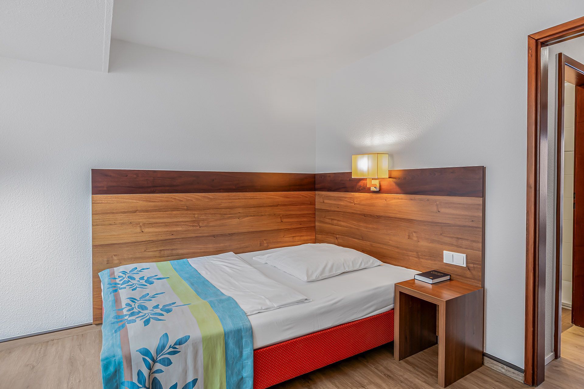 Einzelbett in einem Hotelzimmer mit hölzernem Kopfteil, Nachttisch und offener Tür.