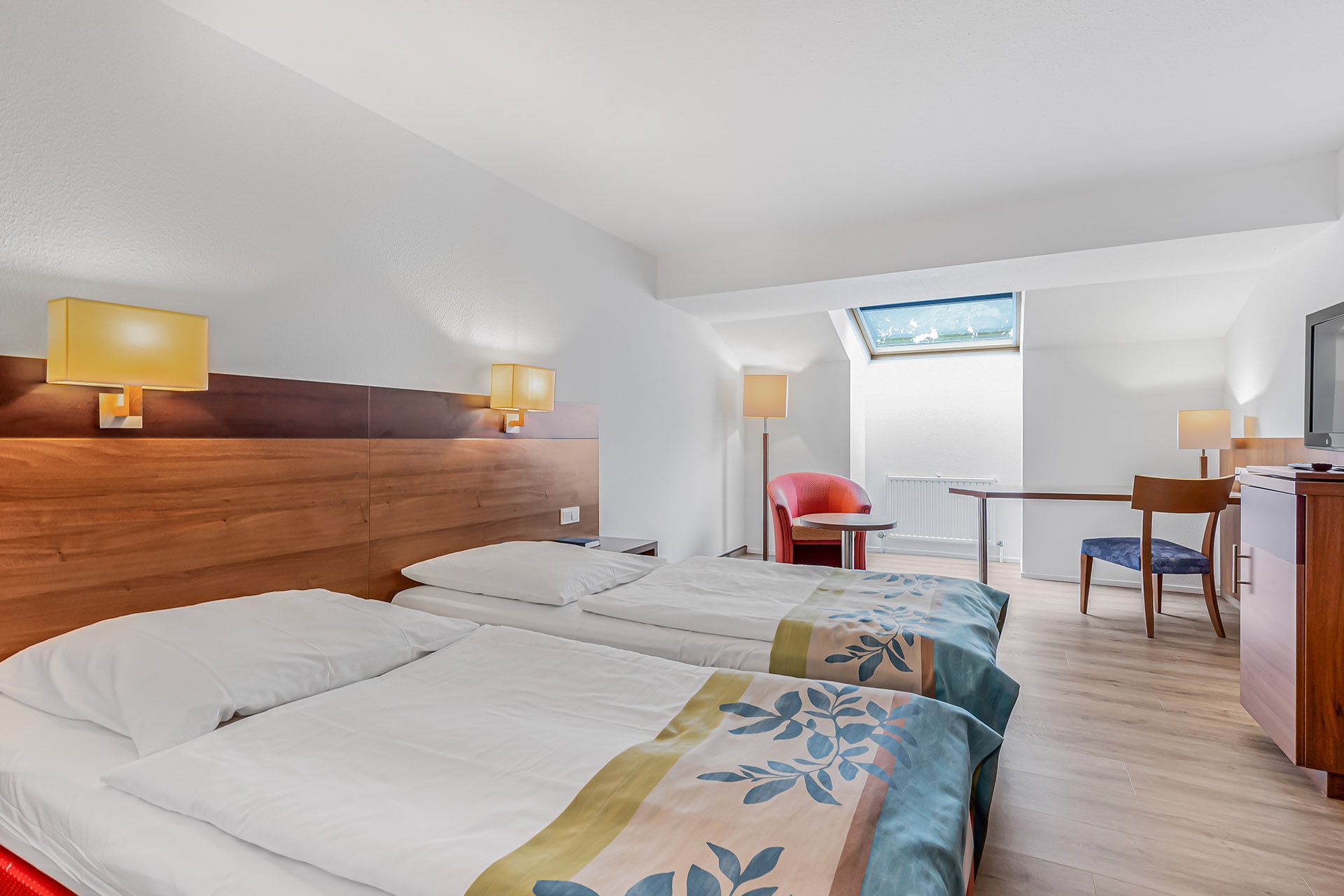 Doppelzimmer mit zwei Betten, Holzkopfteil, Dachfenster, Schreibtisch, Stuhl, rotem Sessel.