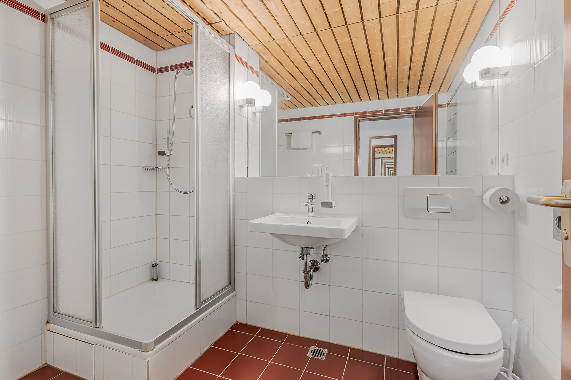 Weißes Badezimmer mit Dusche, Waschbecken und Toilette; brauner Boden und Holzdecke.