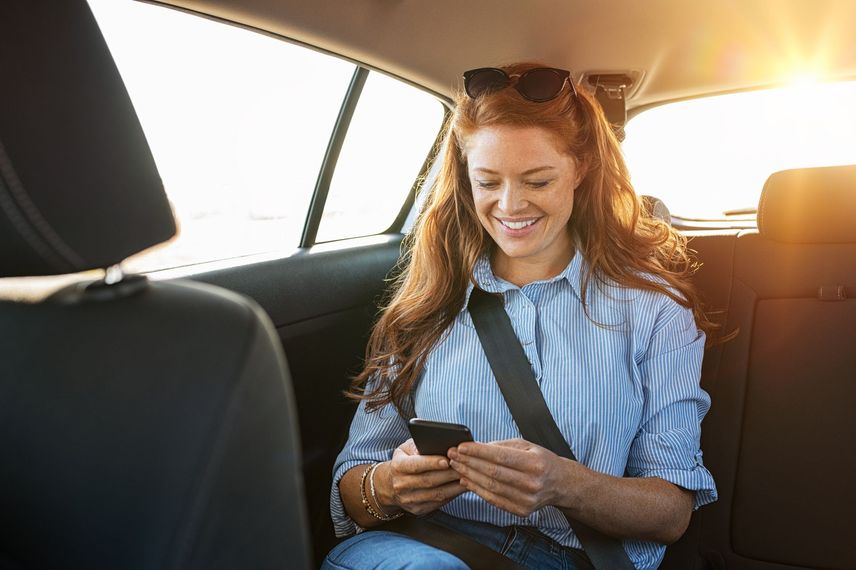 Une femme sourit à son téléphone sur la banquette arrière d'une voiture, la lumière du soleil inondant la pièce par la fenêtre.
