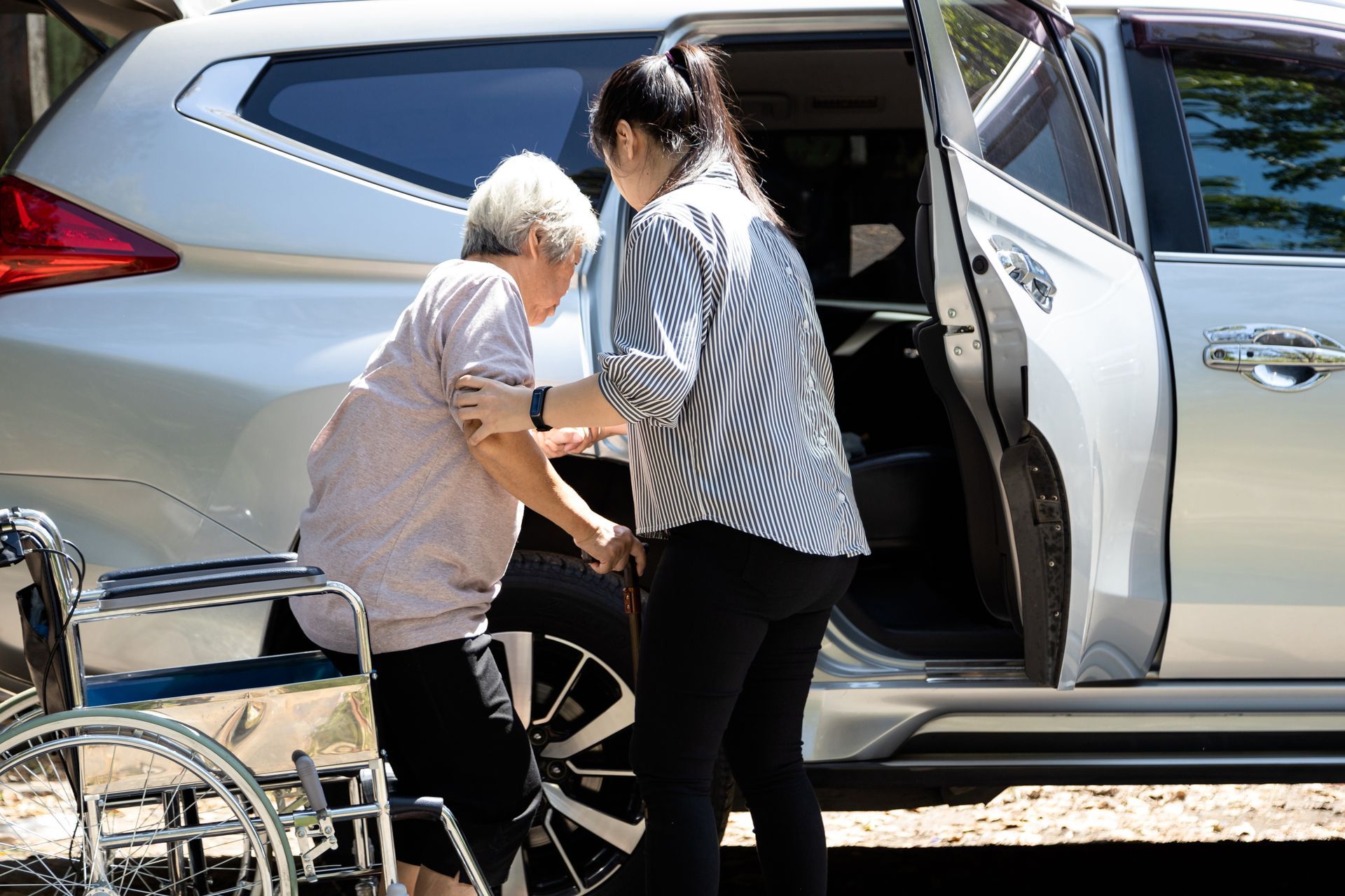 Une femme aide une personne âgée à monter dans une voiture argentée à côté d'un fauteuil roulant.