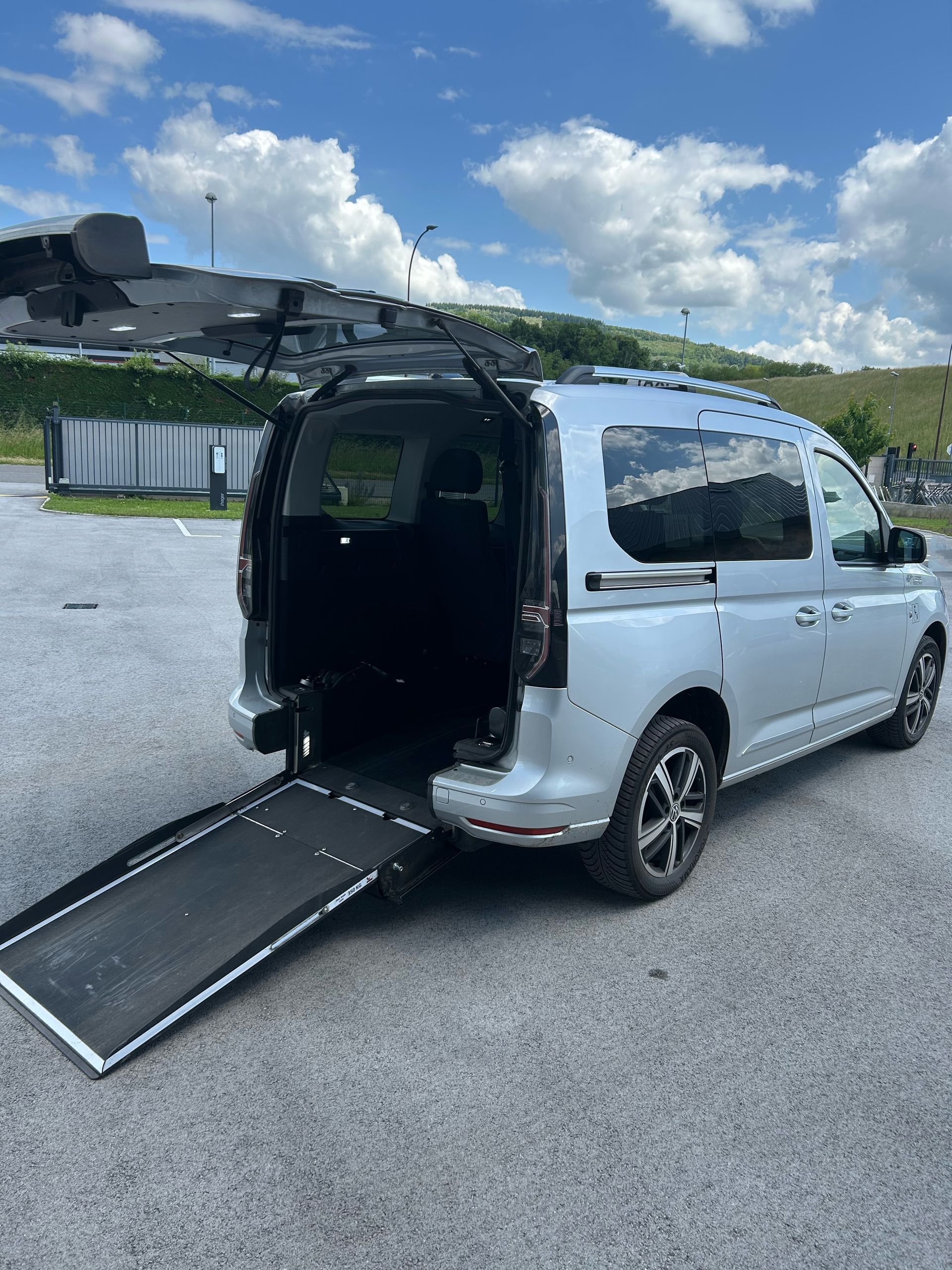 Taxi avec rampe d'accès pour fauteuil roulant déployée à l'arrière, sur un parking sous un ciel nuageux.