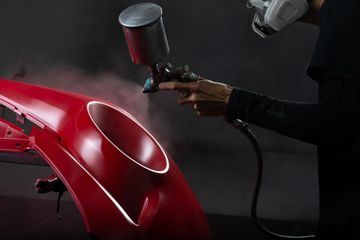 Un ouvrier peint la carrosserie d'une voiture rouge au pistolet à peinture dans un atelier sombre.