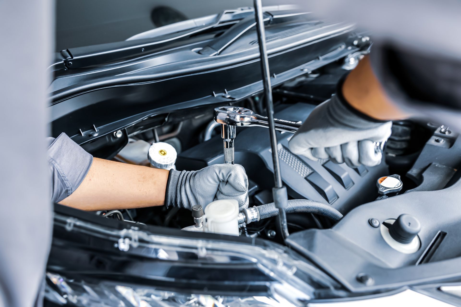 Un mécanicien portant des gants gris inspecte le compartiment moteur d'une voiture, capot ouvert.