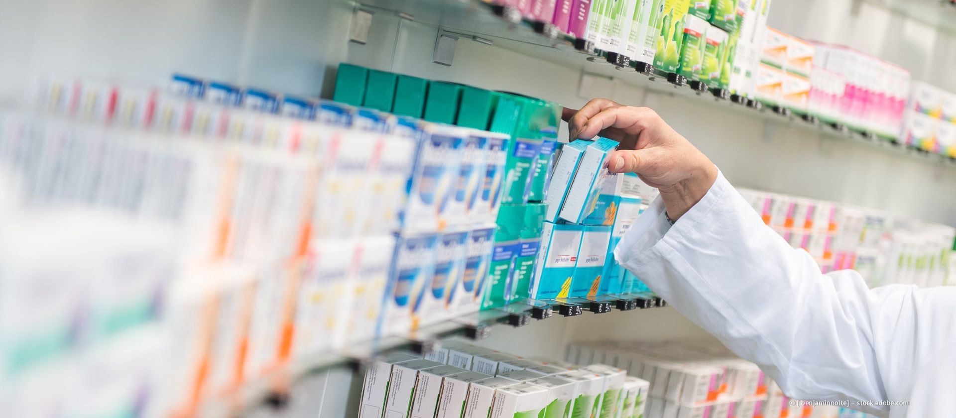 Ein Apotheker im weißen Kittel greift in einer Apotheke nach einer Medikamentenschachtel auf einem Regal.