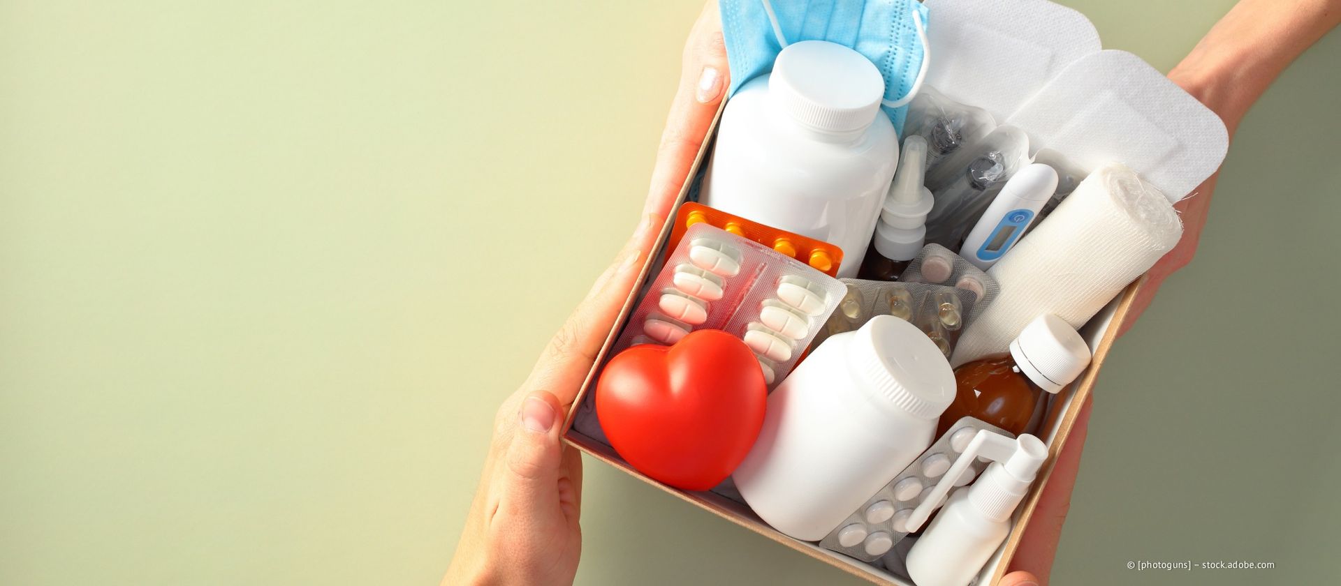 Person hält eine Schachtel mit Medikamenten, einem roten Herzen und einer OP-Maske. Die Schachtel steht vor einem hellgrünen Hintergrund.