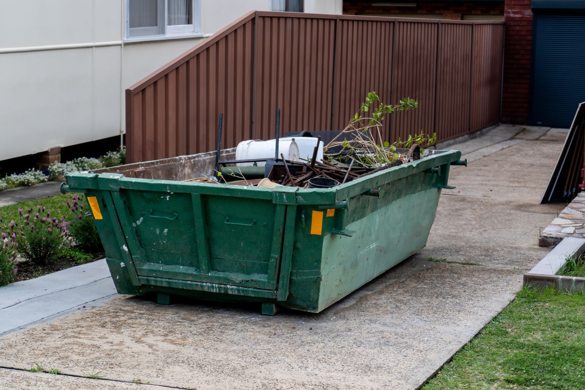 Un conteneur à déchets verts, rempli de déchets verts, est posé sur une allée en béton, à côté d'une maison et d'une clôture.
