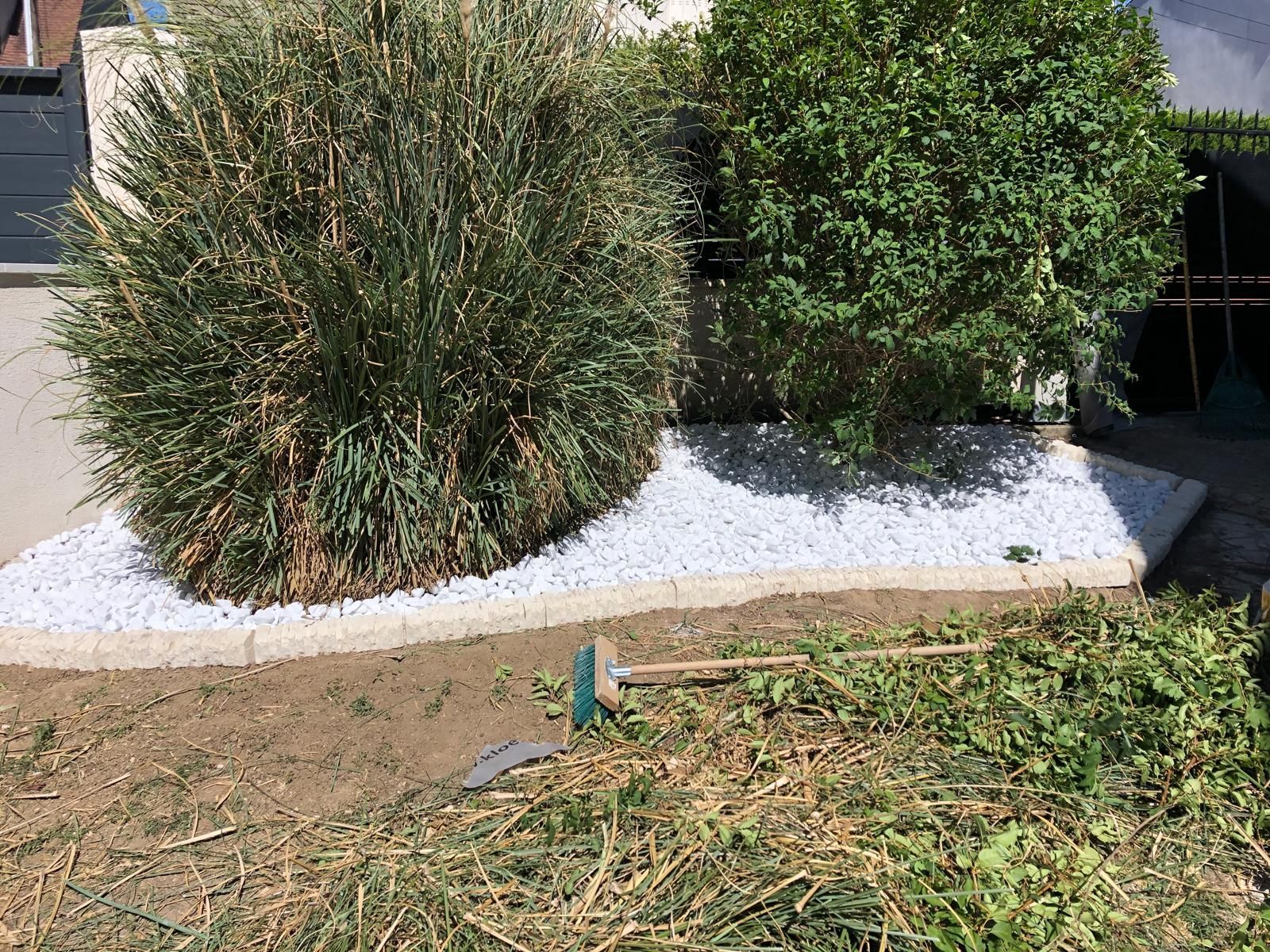 Un parterre de jardin recouvert de gravier blanc, avec deux buissons et un râteau au sol.