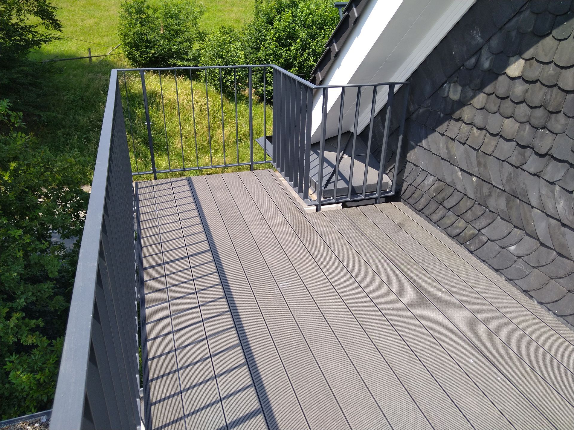 Graue Dachterrasse mit Metallgeländer, Holzdeck und dahinter grünen Bäumen