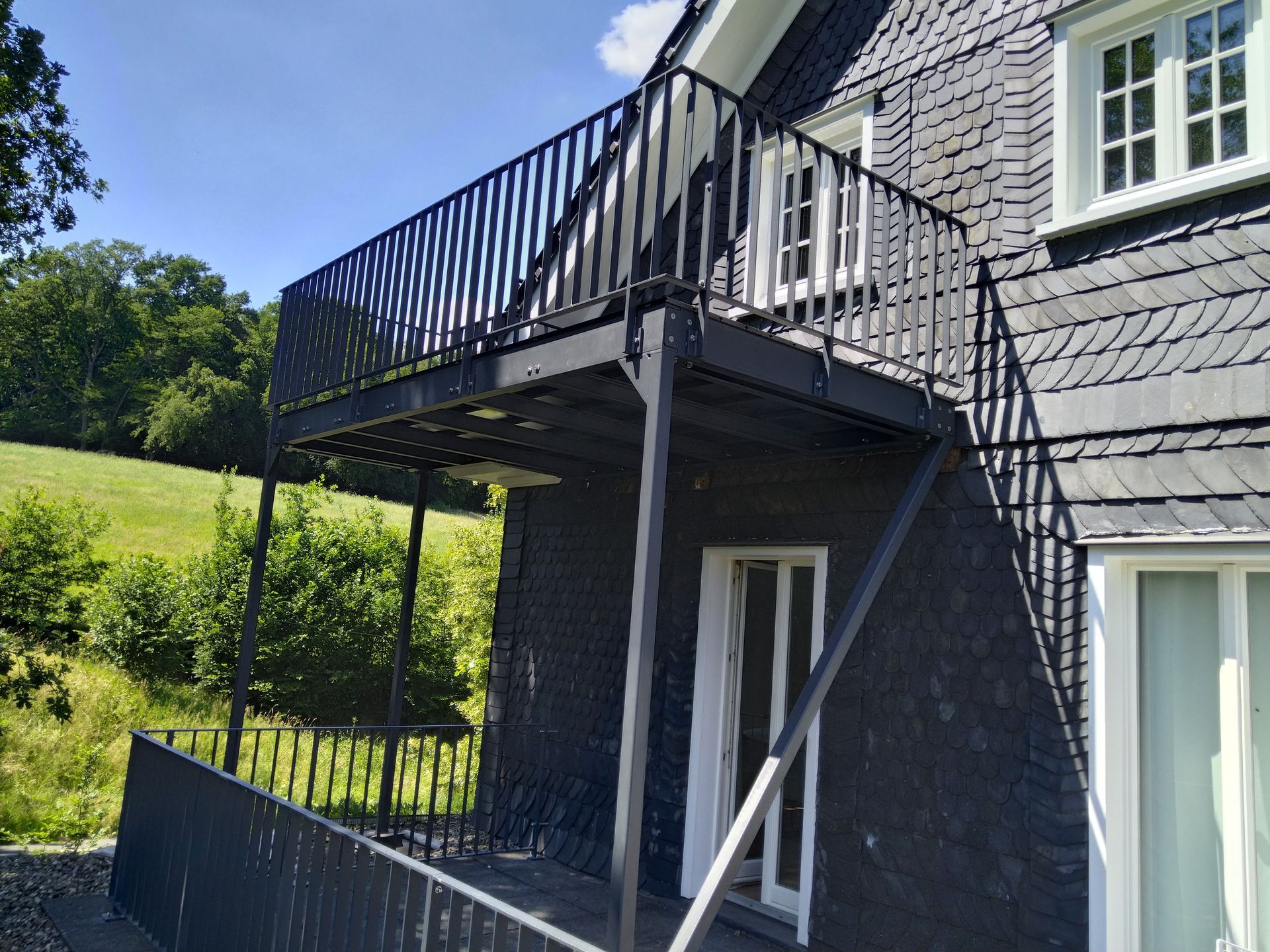 Modernes Haus mit dunkler Fassadenverkleidung und Treppe zum oberen Balkon in einem sonnigen Garten