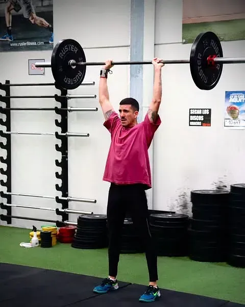 Un hombre con una camisa rosa levanta una barra sobre su cabeza.