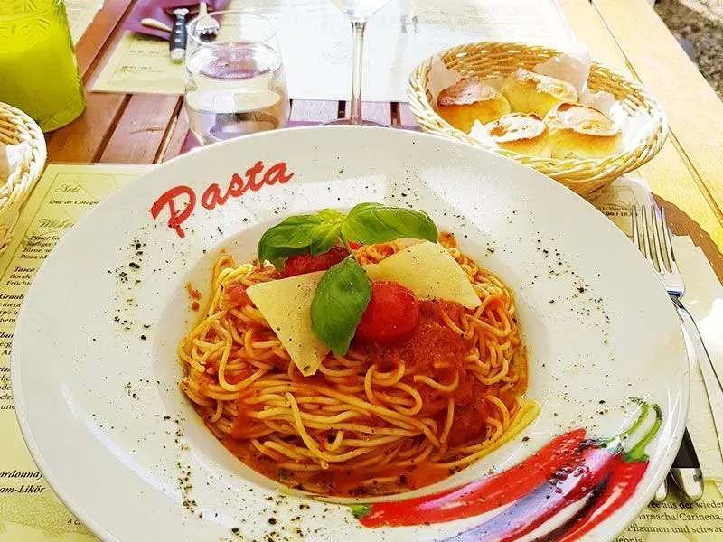 Spaghettigericht mit Tomatensoße, Basilikum und Parmesankäse auf einem weißen Teller an einem Tisch im Freien.