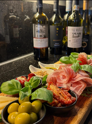 Wurstplatte mit Oliven, Prosciutto, Basilikum und getrockneten Tomaten; im Hintergrund Weinflaschen.