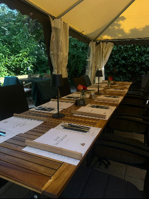 Langer Holztisch zum Essen unter einem cremefarbenen Baldachin, mit Tischsets, Besteck und schwarzen Stühlen.