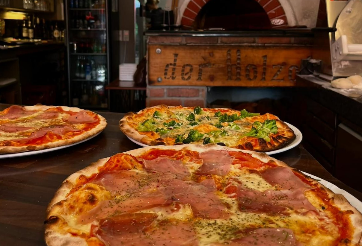 Drei Pizzen auf einem Tisch in einem Restaurant. Mit Schinken und Brokkoli belegt, im Hintergrund ein Steinofen.