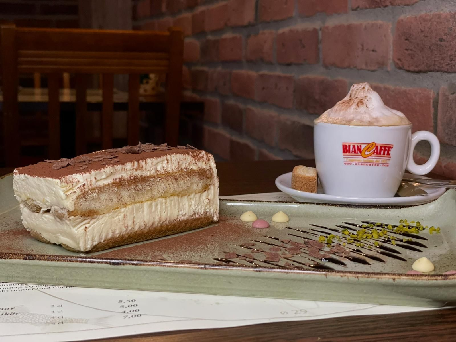 Tiramisu-Dessert mit Cappuccino, serviert auf einem Teller in einem Café mit Backsteinwänden.