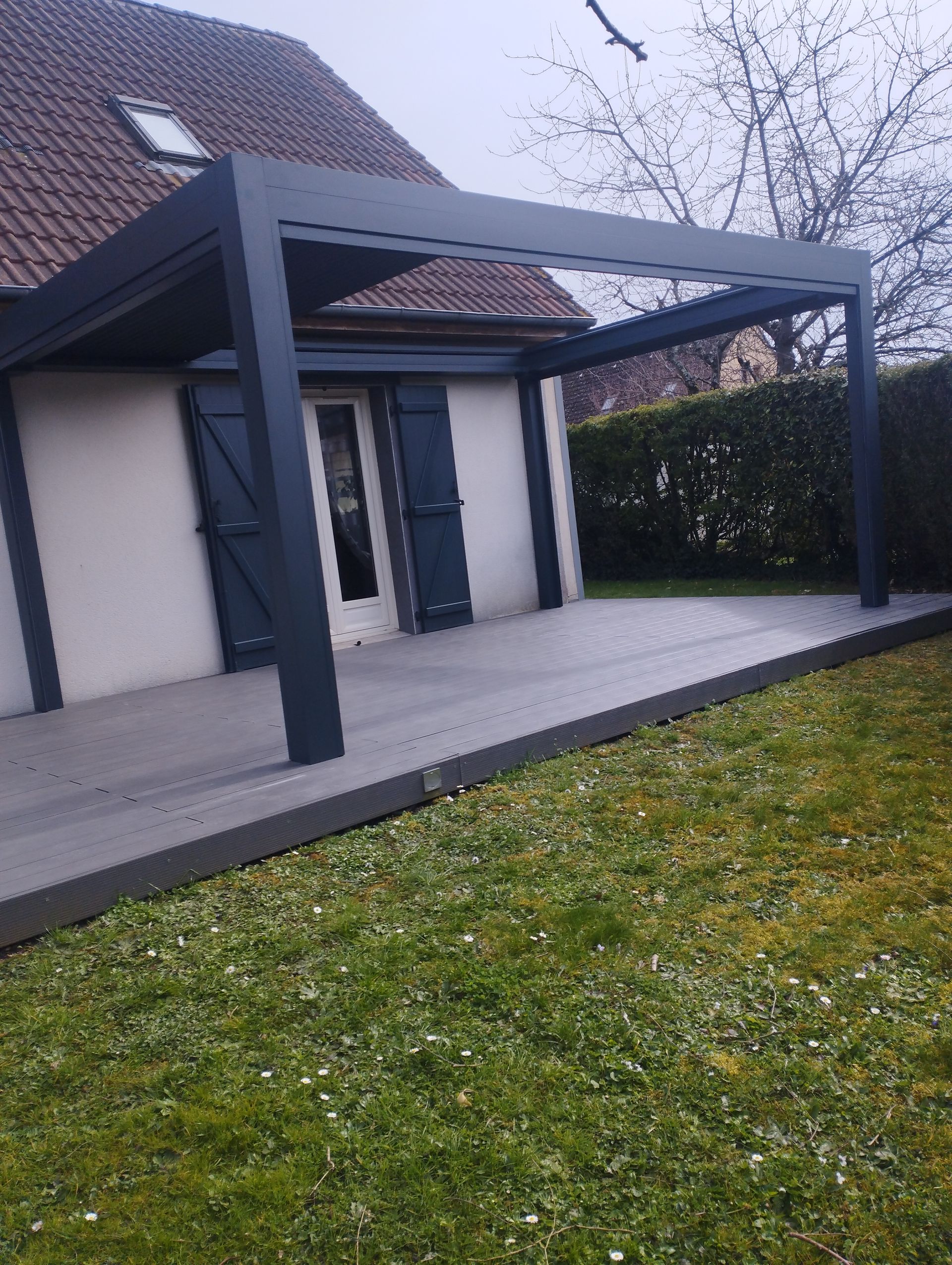 Pergola sur une terrasse jouxtant une maison à volets gris foncé