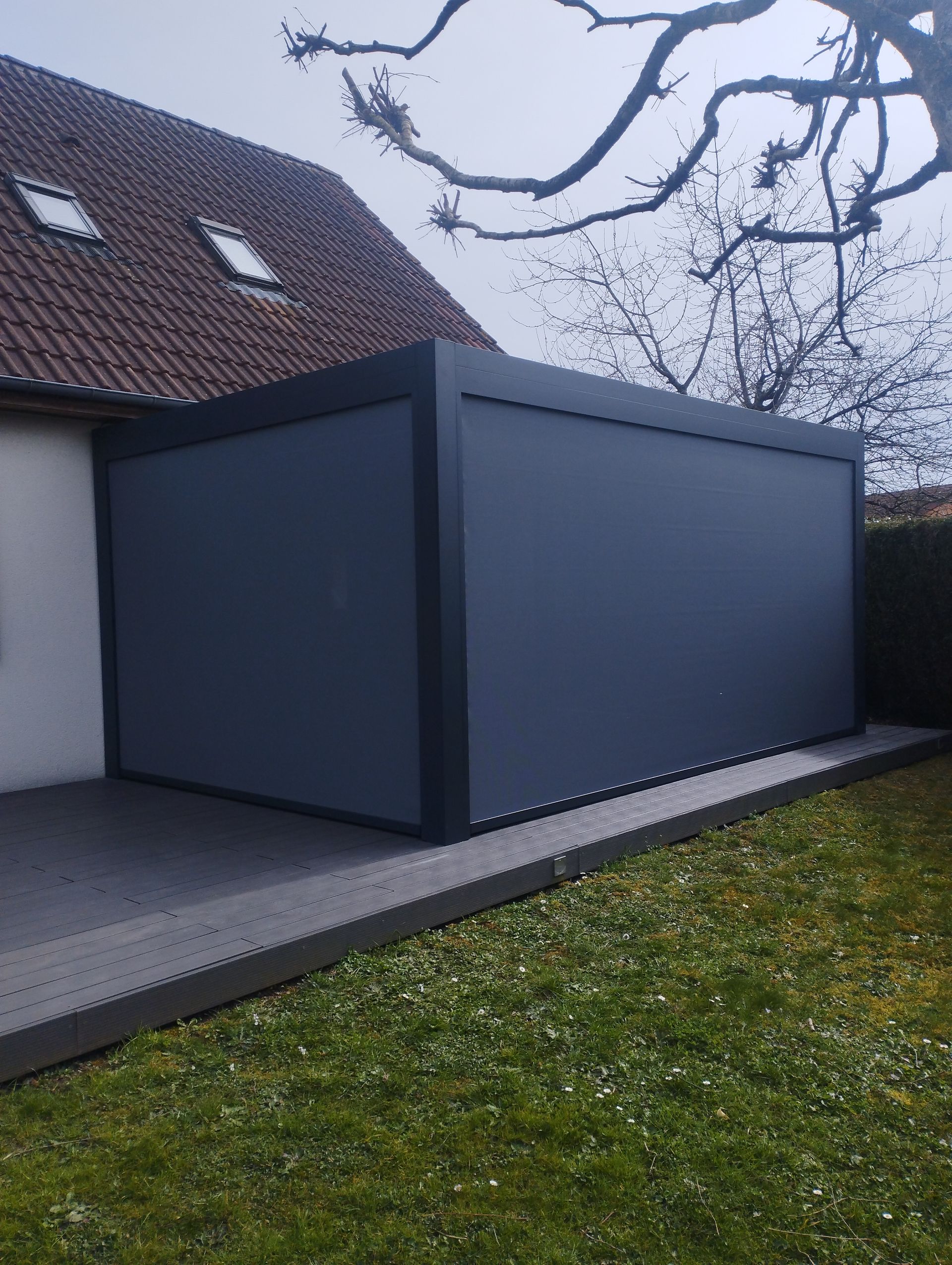 Pergola grise avec panneaux rétractables, adjacente à une maison, sur une terrasse en béton