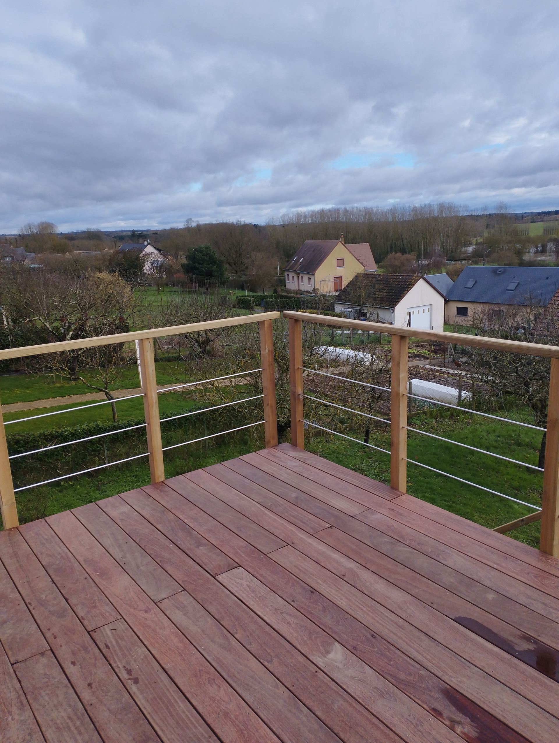 Terrasse en bois avec rambarde en câbles donnant sur un paysage rural avec des maisons et des arbres