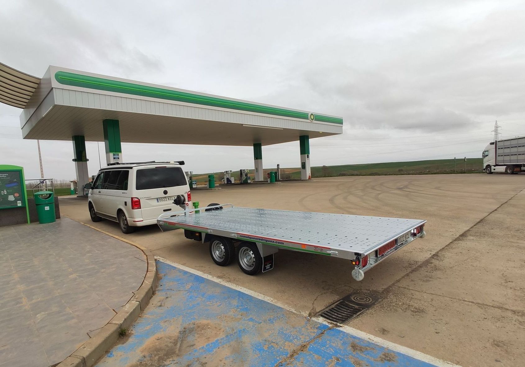 Una camioneta blanca con remolque de plataforma en una gasolinera, bajo una marquesina verde y blanca. Cielo nublado.