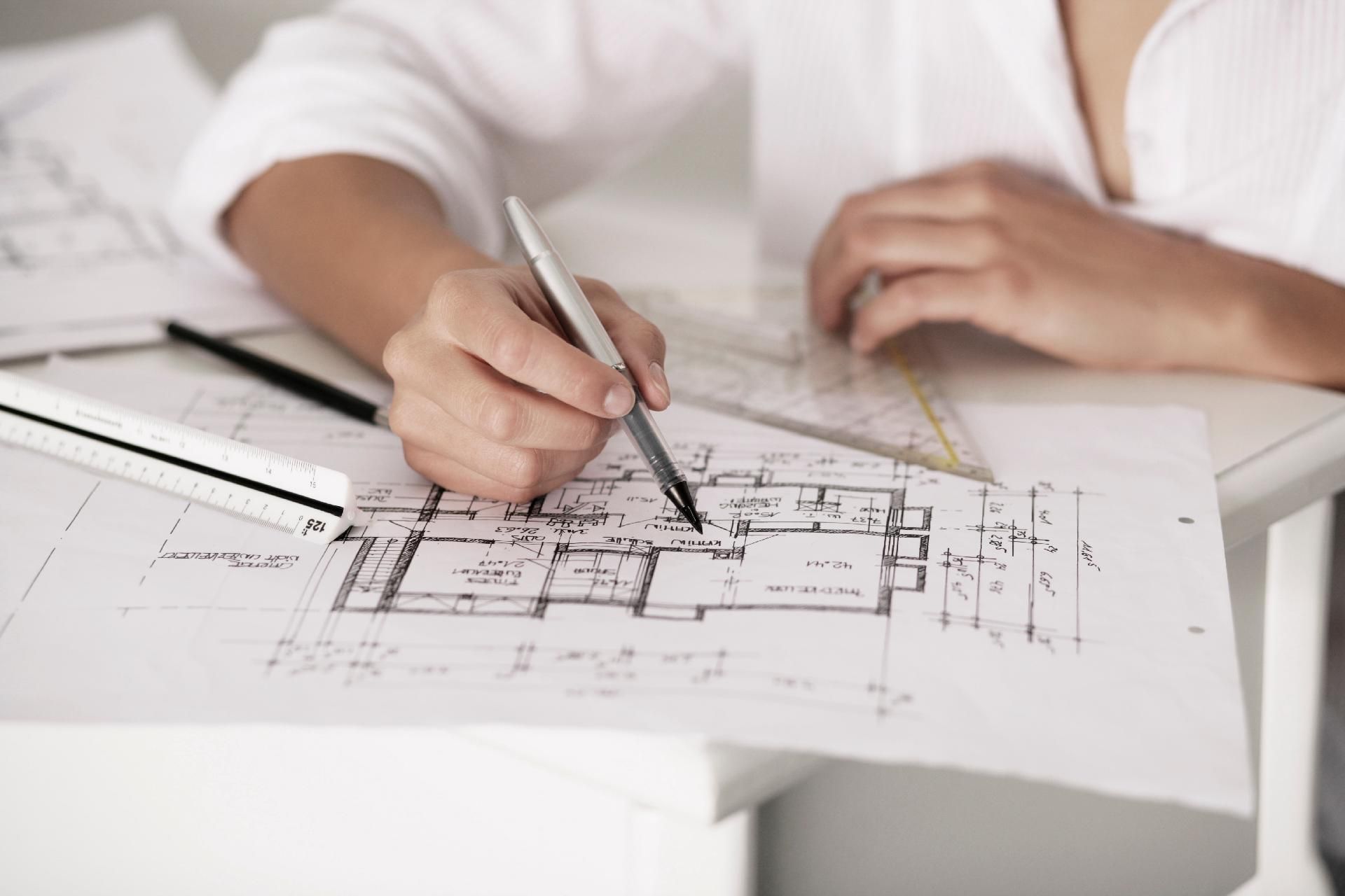 Eine Person zeichnet mit einem Stift und einem Geodreieck Details in einen technischen Bauplan auf einem Schreibtisch.