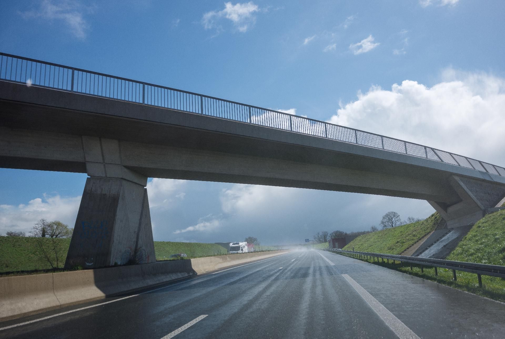 Eine moderne Betonbrücke führt über eine mehrspurige Autobahn unter einem leicht bewölkten blauen Himmel.