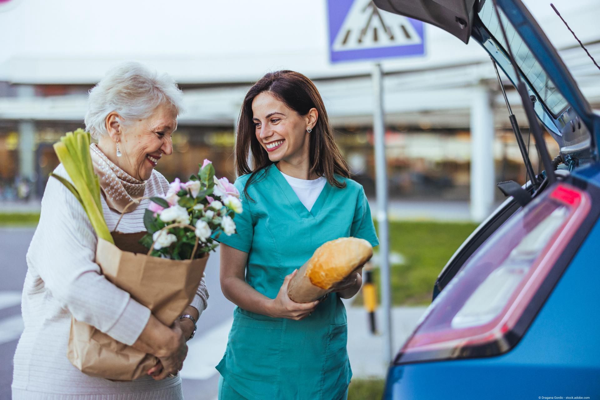 Eine junge Frau in blauer Arbeitskleidung hilft einer Seniorin beim Einladen von Einkaufstüten und Blumen in ein Auto.