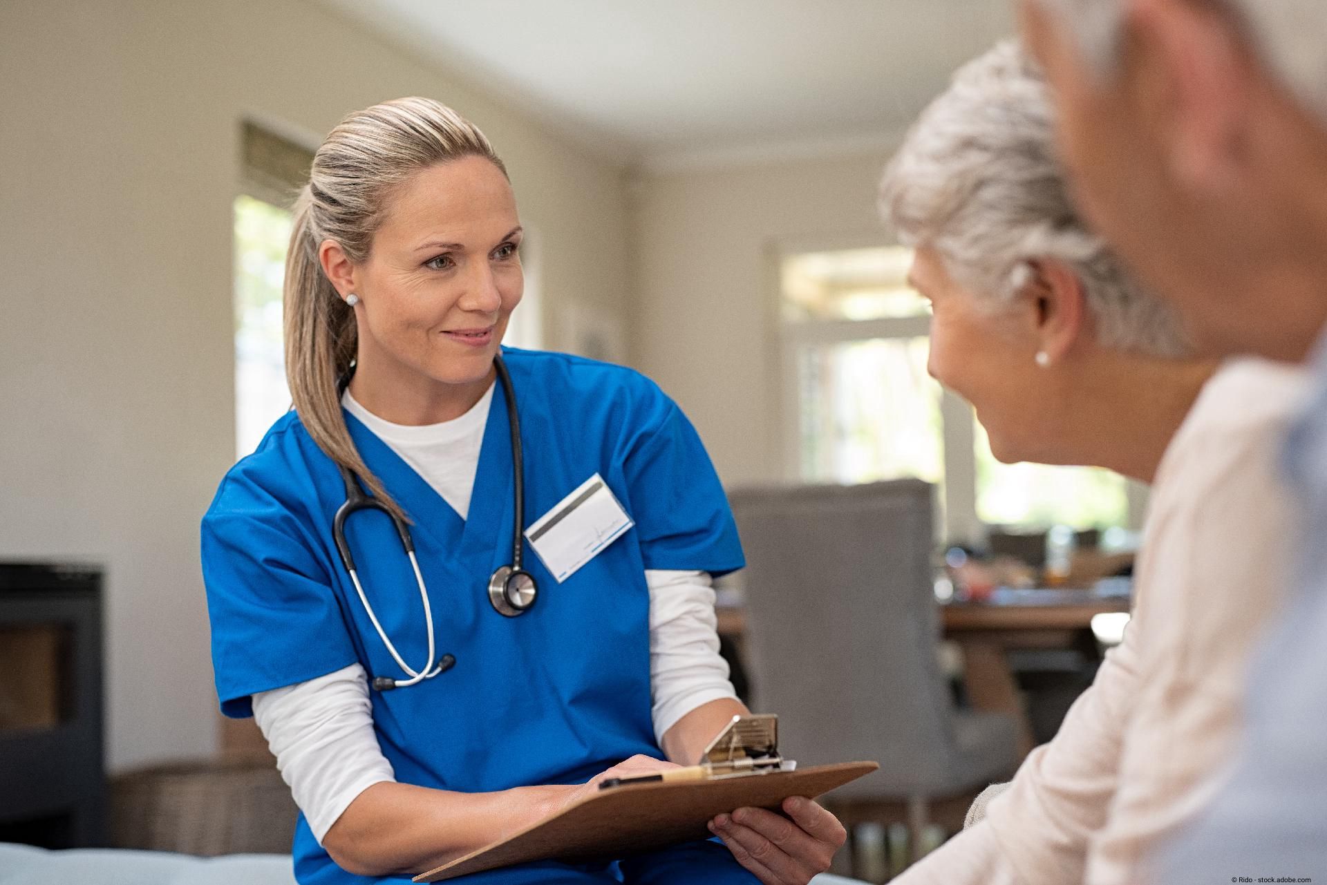 Eine Krankenschwester in blauer Uniform sitzt mit einem Klemmbrett im Gespräch einer älteren Person gegenüber.