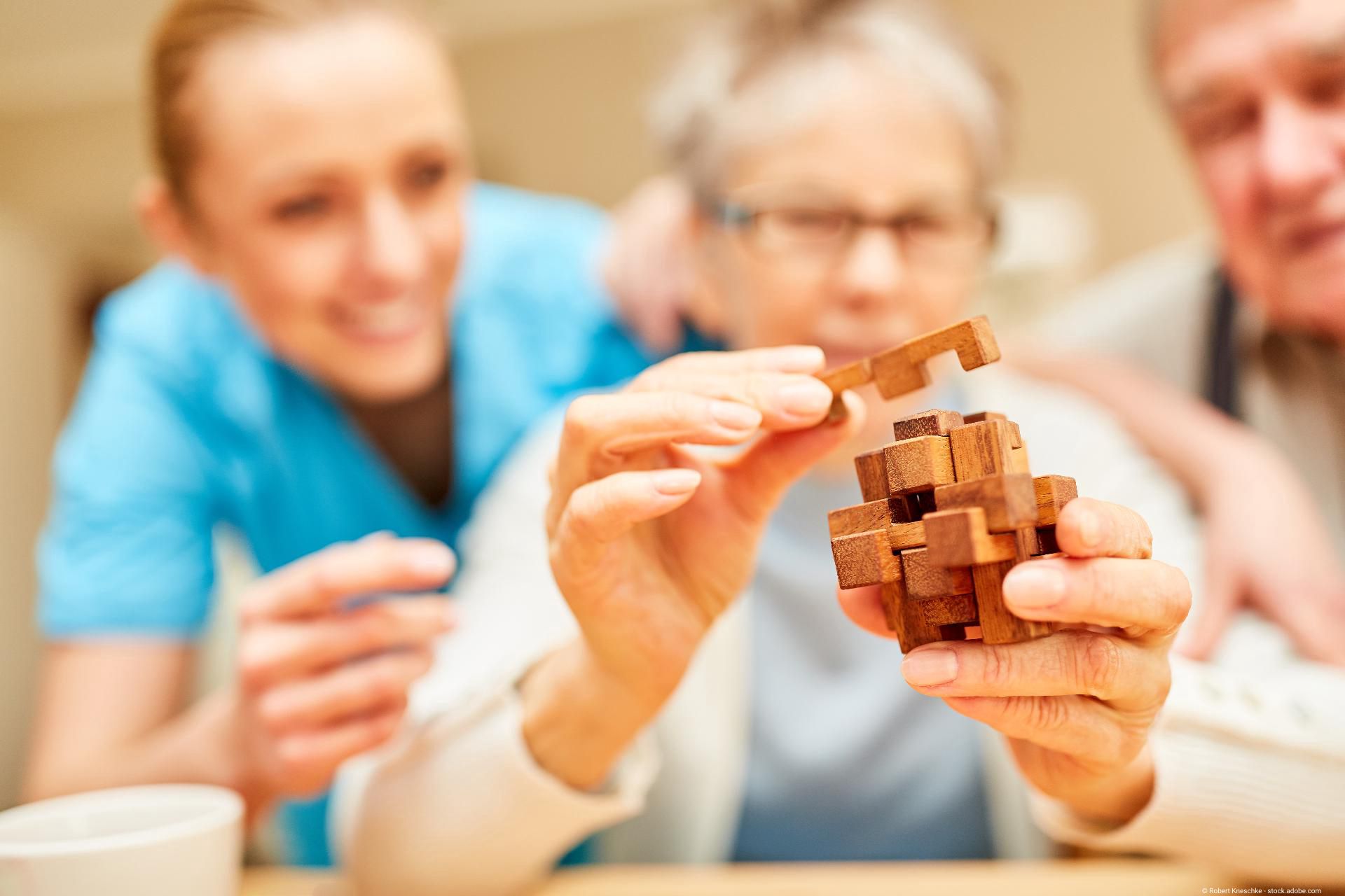 Eine Pflegekraft beobachtet lächelnd, wie eine ältere Frau konzentriert an einem hölzernen 3D-Puzzle arbeitet.