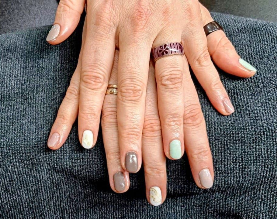 Manos con uñas pintadas en varios colores neutros, anillos en los dedos, apoyadas sobre una superficie azul.