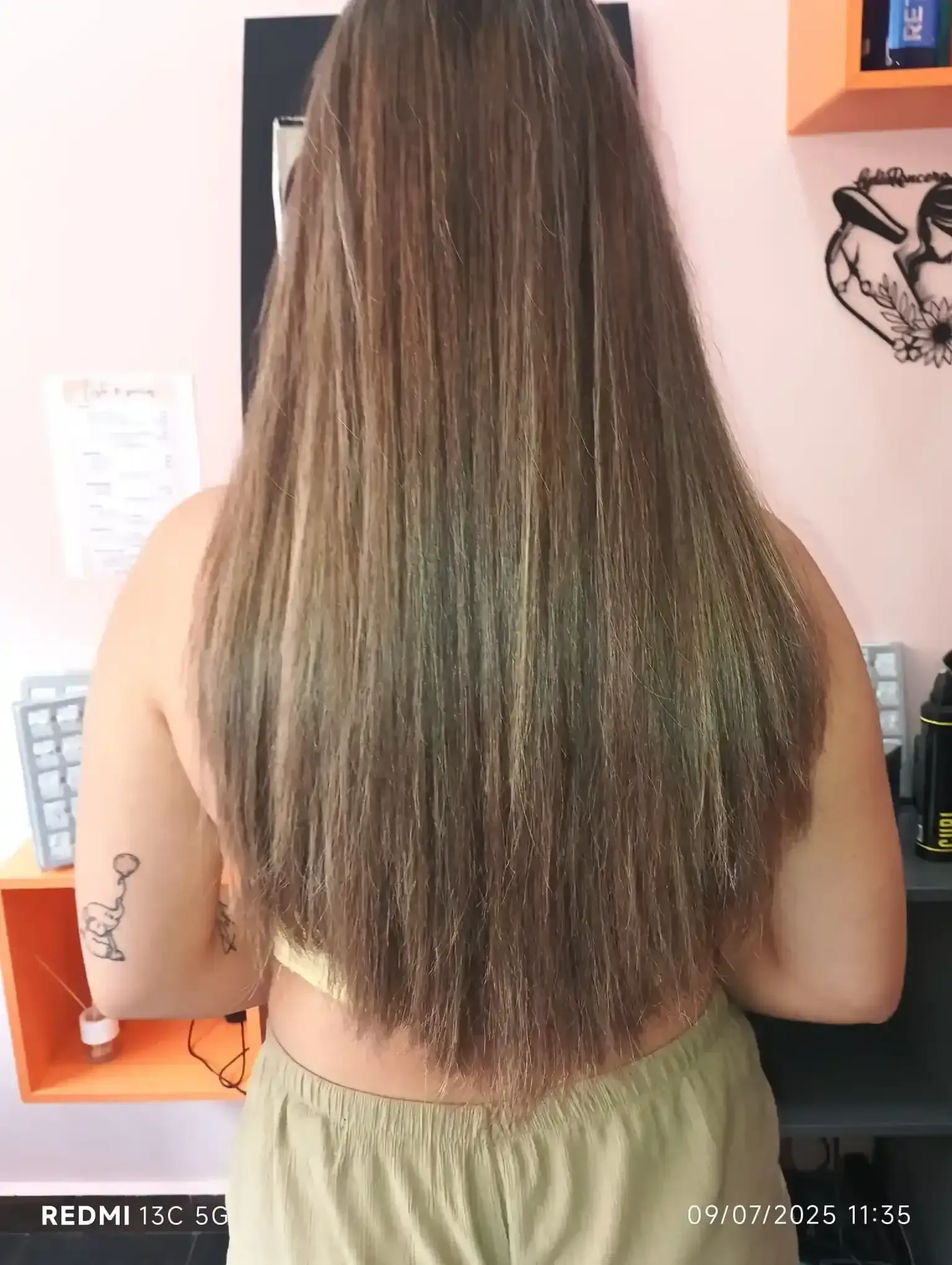 Persona con cabello largo y liso con reflejos, de pie en interiores con un escritorio y una pared rosa al fondo.