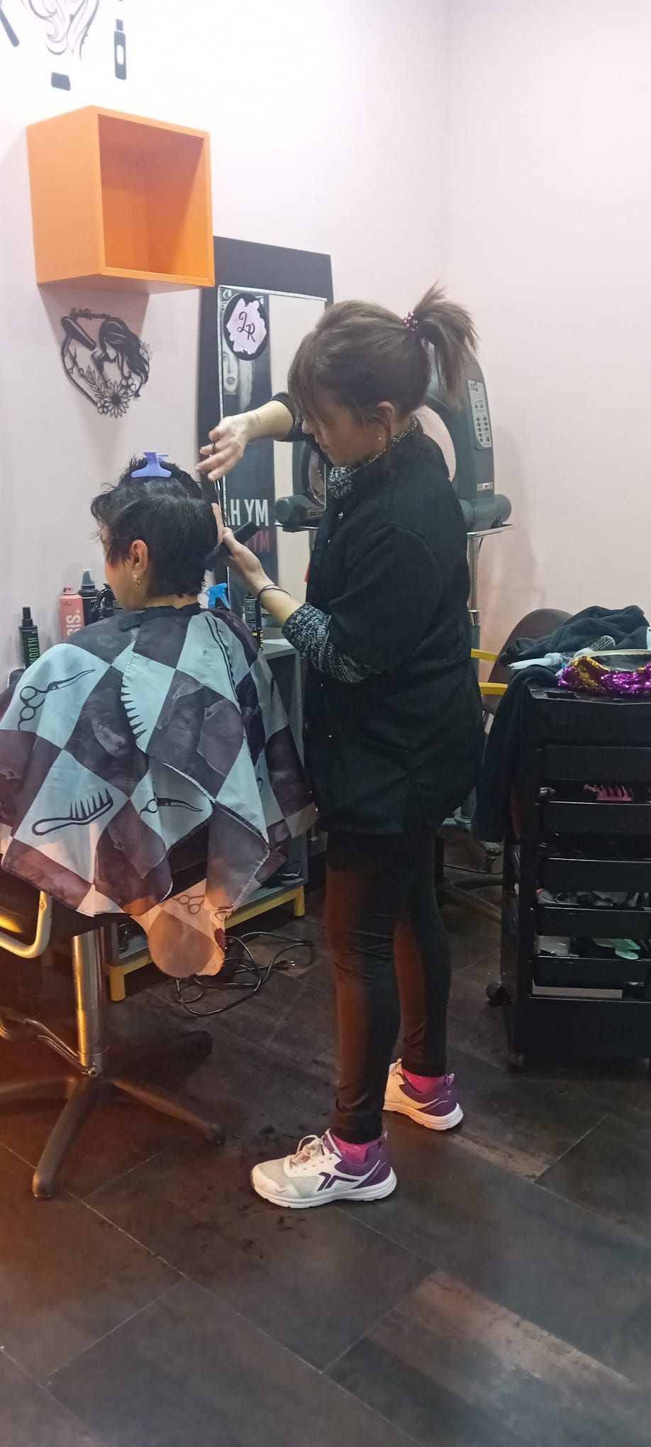 Una persona en una peluquería recibiendo un corte de pelo por parte de un estilista. Suelo de madera oscura, estante naranja en la pared y capa negra y plateada.