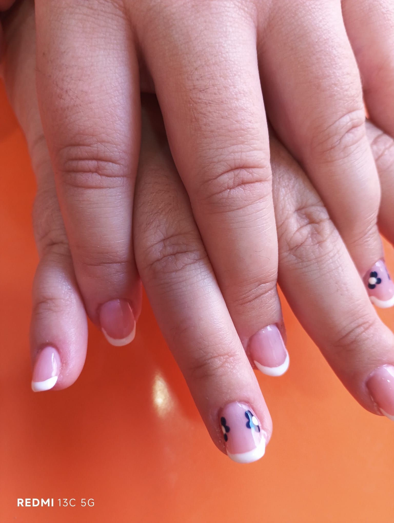 Manos con uñas con manicura francesa, una con un pequeño diseño floral, sobre una superficie naranja.