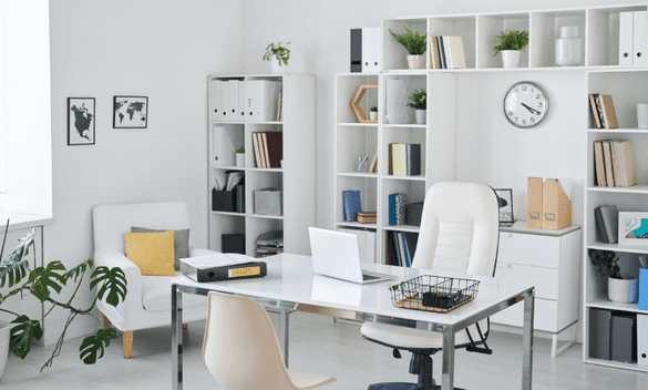 Oficina moderna y luminosa con estanterías blancas, escritorio, sillas, plantas y un reloj de pared.