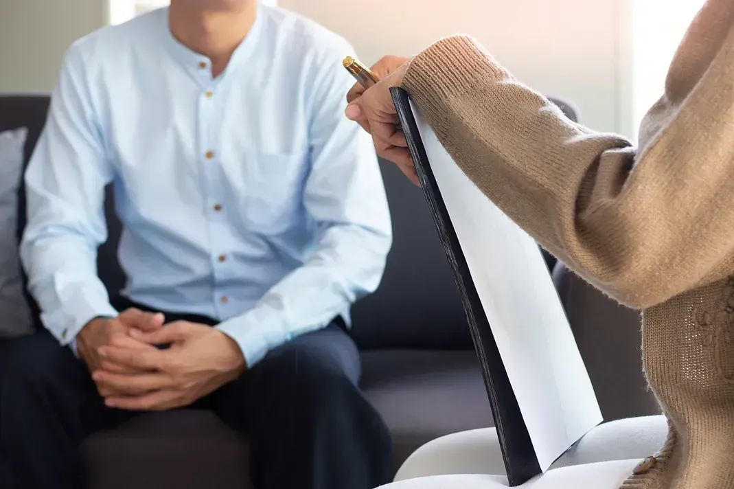 Dos personas en una sesión de terapia, una sosteniendo un portapapeles y la otra sentada con las manos entrelazadas.