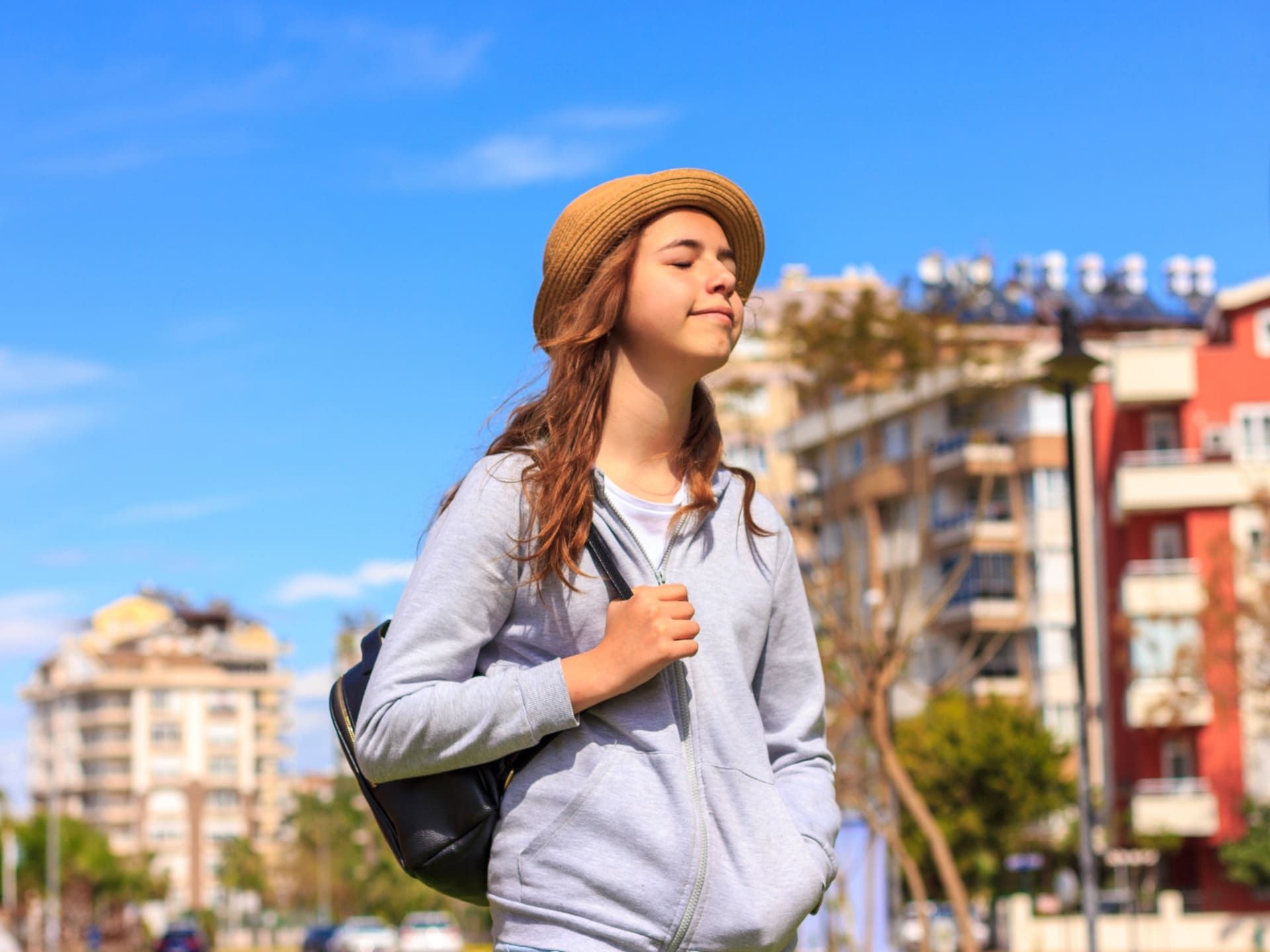 Mujer con sombrero de paja caminando al aire libre, sonriendo bajo un cielo azul con edificios al fondo.