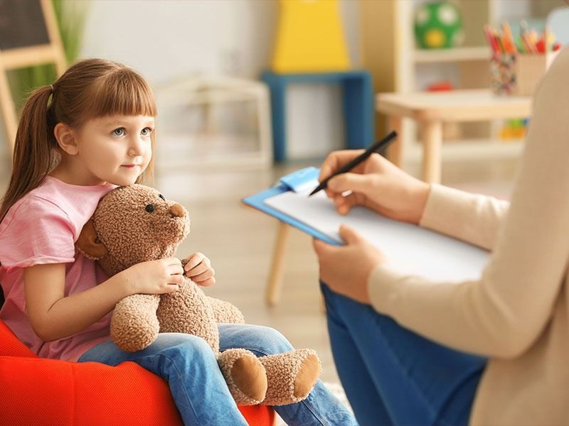 Un niño que sostiene un osito de peluche habla con un consejero durante una sesión de terapia en una luminosa sala de juegos.