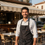 Empleado de cafetería sonriente con un delantal negro, de pie frente a un restaurante con mesas y toldo