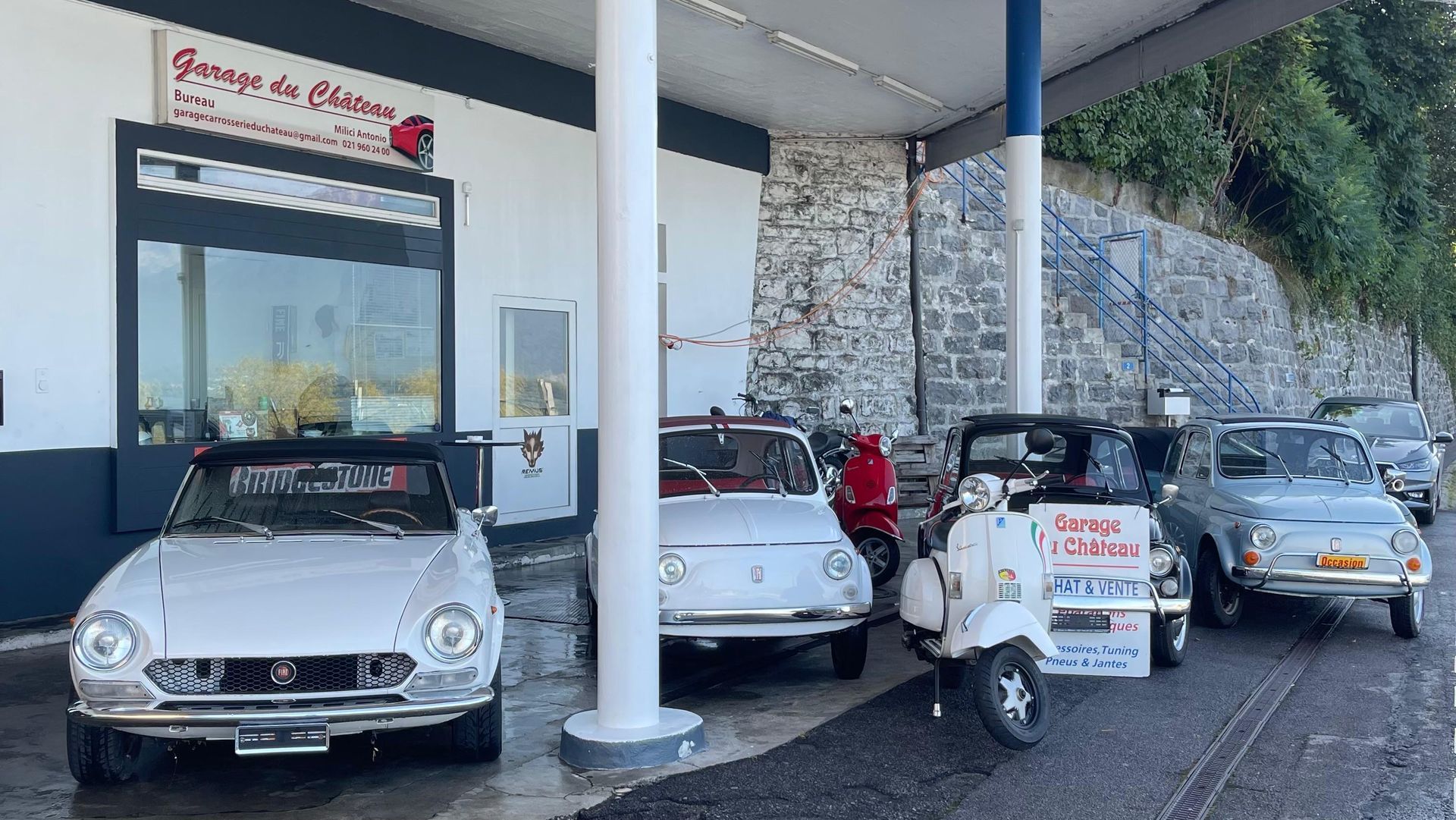 Voitures classiques et scooter garés dans un garage avec un panneau.