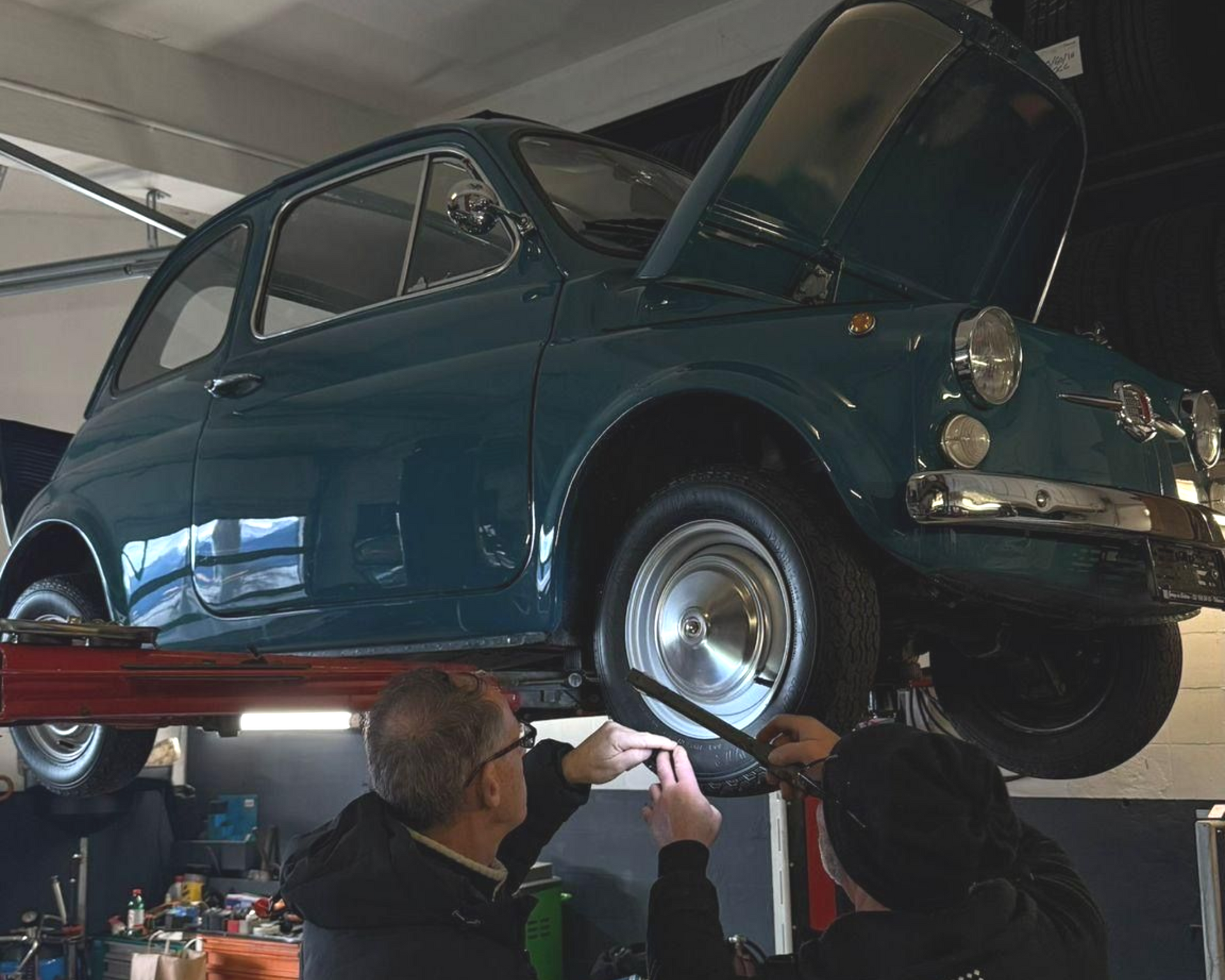 Fiat 500 bleu sarcelle sur un ascenseur, avec deux personnes inspectant une roue dans un garage.