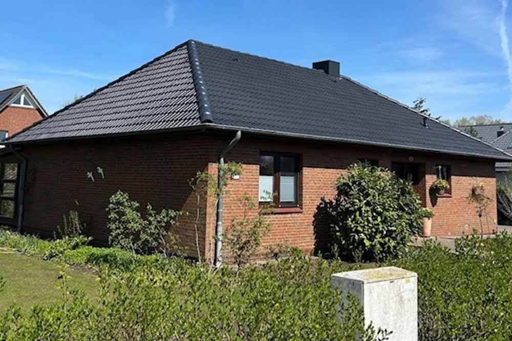 Ein Backsteinhaus mit schwarzem Dach ist von Büschen und Bäumen umgeben.